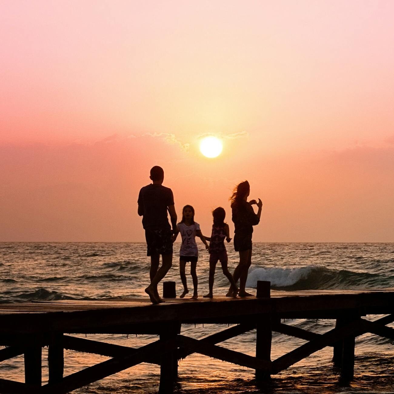 Silhouetted family enjoys a stroll on the beach pier at a vibrant sunset over the ocean waves.